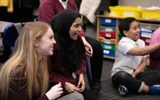 A teacher and students are sitting on the floor of a classroom. They are smiling and laughing.