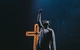 An actor is standing in front of a glowing cross. They are gesturing upwards.
