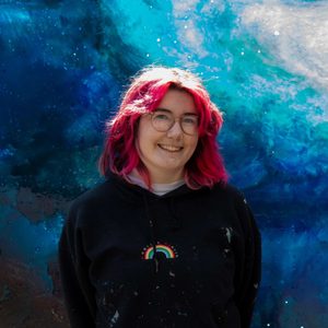 Eilidh is smiling at the camera. She has pink hair and is wearing glasses.