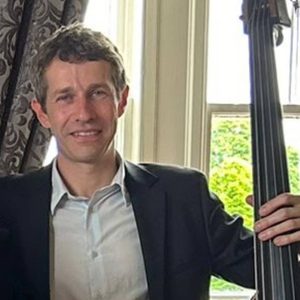 Tom is smiling at the camera. He is holding a double bass.