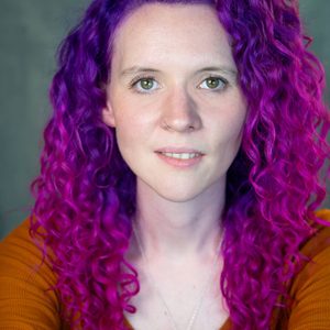 Fiona is looking at the camera. She has purple and pink hair.